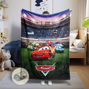May include: A plush blanket featuring the animated character Lightning McQueen from the movie Cars. The blanket showcases a race scene with other Cars characters on a green field, with a stadium backdrop. The blanket has a red, white, and black logo that says "Name".