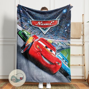 May include: A plush blanket featuring a vibrant image of Lightning McQueen from the Cars movie. The blanket showcases a race scene with the Cars logo and the name "Name" in a stylized font. The primary colors are red, blue, and green.