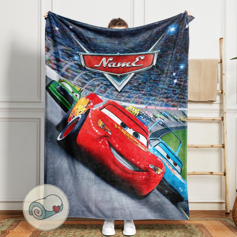 May include: A plush blanket featuring a vibrant image of Lightning McQueen from the Cars movie. The blanket showcases a race scene with the Cars logo and the name "Name" in a stylized font. The primary colors are red, blue, and green.