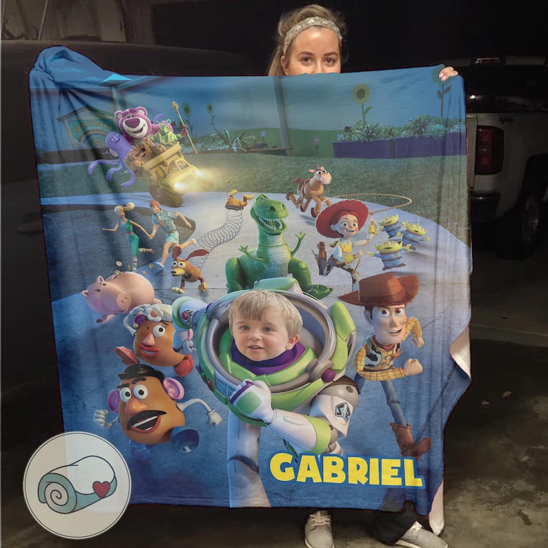 May include: A blue blanket featuring characters from the Toy Story movie, including Buzz Lightyear, Woody, and Rex. The blanket has a personalized name, "GABRIEL", printed in yellow. The blanket also features a picture of a child.