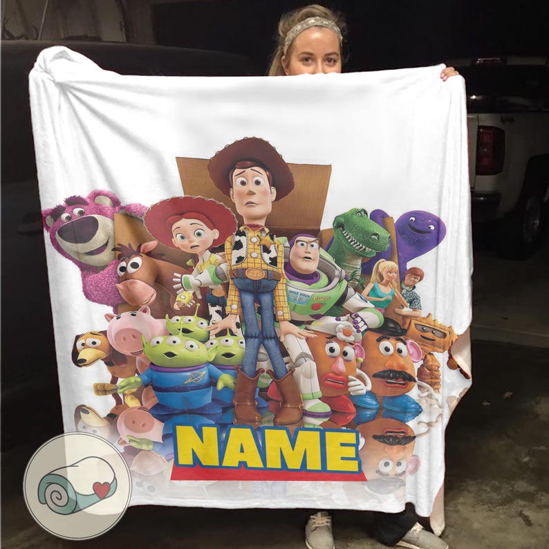 May include: White blanket with a colourful graphic of Toy Story characters, including Woody, Buzz Lightyear, and others. The blanket features the word "NAME" in yellow and blue. The blanket is likely made of soft material.