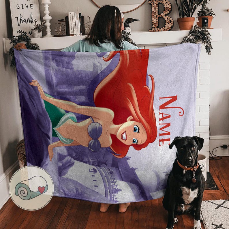 May include: A soft, purple blanket featuring a cartoon illustration of a mermaid with red hair, a blue bra, and a green tail. The blanket has the word "NAME" in red script. A black dog with a brown collar sits nearby.