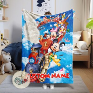 May include: A blue blanket featuring a colorful illustration of Mickey Mouse, Minnie Mouse, and other Disney characters on a train. The blanket has the text "CUSTOM NAME" in red. The background is a blue sky with clouds.