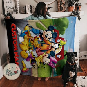 K&ouml;nnte beinhalten: Eine farbenfrohe Decke mit Disney-Figuren wie Micky Maus, Donald Duck und Minnie Maus. Die Decke hat einen individuellen Namen an der Seite und eine Grafik einer zusammengerollten Decke. Ein schwarzer Hund mit Halsband sitzt vor der Decke.