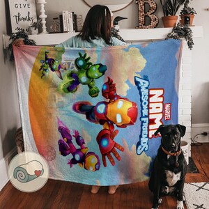 K&ouml;nnte beinhalten: Eine Marvel "Awesome Friends" Decke mit Iron Man und anderen Charakteren in einem farbenfrohen Design. Die Decke hat einen blauen Hintergrund mit dem Marvel-Logo und dem Wort "NAME" darauf. Ein schwarzer Hund sitzt auf dem Boden.