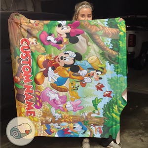 May include: A colorful blanket featuring Mickey Mouse, Minnie Mouse, Goofy, Donald Duck, and other cartoon characters in a forest setting. The blanket has the words "CUSTOM NAME" printed vertically in red. The blanket is held up by a person.