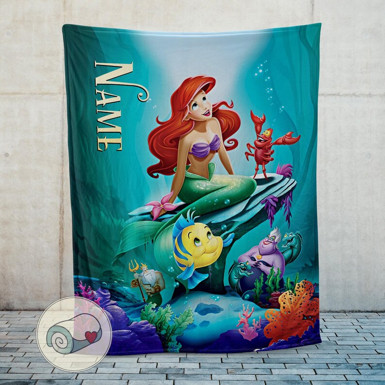 Puede incluir: Una manta turquesa con una colorida ilustraci&oacute;n de Ariel de La Sirenita. Ariel, con cabello rojo y cola verde, est&aacute; sentada en una roca con Flounder, Sebasti&aacute;n y &Uacute;rsula. La palabra "NAME" est&aacute; impresa verticalmente.