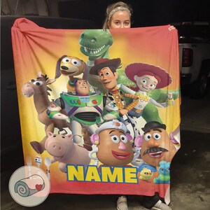 May include: A large, rectangular blanket featuring characters from the animated film Toy Story. The blanket has a vibrant design with Woody, Buzz Lightyear, and other characters. The word "NAME" is printed in large yellow letters.