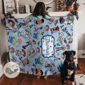 May include: A light blue blanket featuring various Toy Story characters. The blanket has a personalized name tag that says "Thomas". The blanket is decorated with images of Woody, Buzz Lightyear, and other characters from the movie.