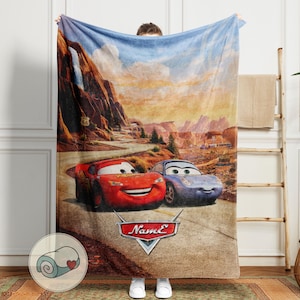 May include: A plush blanket featuring Lightning McQueen and Sally Carrera from the Cars movie, set against a desert landscape with Route 66 signage. The blanket has a red, blue, and brown color scheme, with the Cars logo at the bottom.