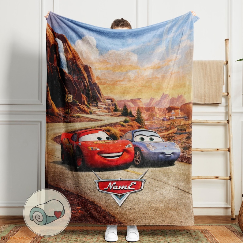 May include: A plush blanket featuring Lightning McQueen and Sally Carrera from the Cars movie, set against a desert landscape with Route 66 signage. The blanket has a red, blue, and brown color scheme, with the Cars logo at the bottom.