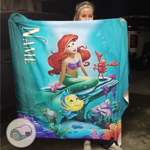 Puede incluir: Una manta turquesa con una colorida ilustraci&oacute;n de Ariel, la sirena de pelo rojo y top morado, sentada en una roca con Flounder y Sebasti&aacute;n. La palabra "NAME" est&aacute; impresa verticalmente en la manta.