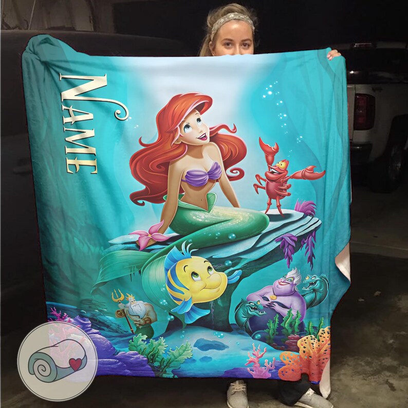 Puede incluir: Una manta turquesa con una colorida ilustraci&oacute;n de Ariel, la sirena de pelo rojo y top morado, sentada en una roca con Flounder y Sebasti&aacute;n. La palabra "NAME" est&aacute; impresa verticalmente en la manta.