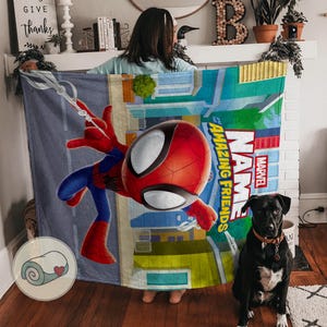 May include: A colorful blanket featuring the Marvel character Spider-Man. The design includes a large image of Spider-Man in action, with the words "Name" and "Amazing Friends" in bold letters. The blanket has a cityscape background.