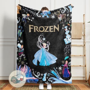 Op de afbeelding: Zwarte fleece deken met personages en het woord "FROZEN" uit de animatiefilm. De deken heeft afbeeldingen van Elsa, Anna, Olaf en andere personages, met sneeuwvlokken en winterlandschap.