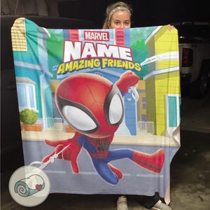 May include: A large, rectangular blanket featuring a cartoon Spider-Man design. The blanket has a cityscape background with the words "MARVEL NAME and his AMAZING FRIENDS" printed across the top. The blanket is primarily blue, red, and yellow.