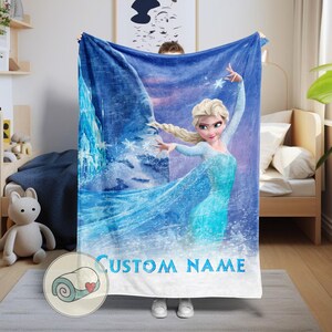 Puede incluir: Una manta polar azul con un dise&ntilde;o de una princesa de dibujos animados con un vestido azul, con el texto "CUSTOM NAME". La princesa est&aacute; situada sobre un fondo de hielo y nieve, con un cielo estrellado.