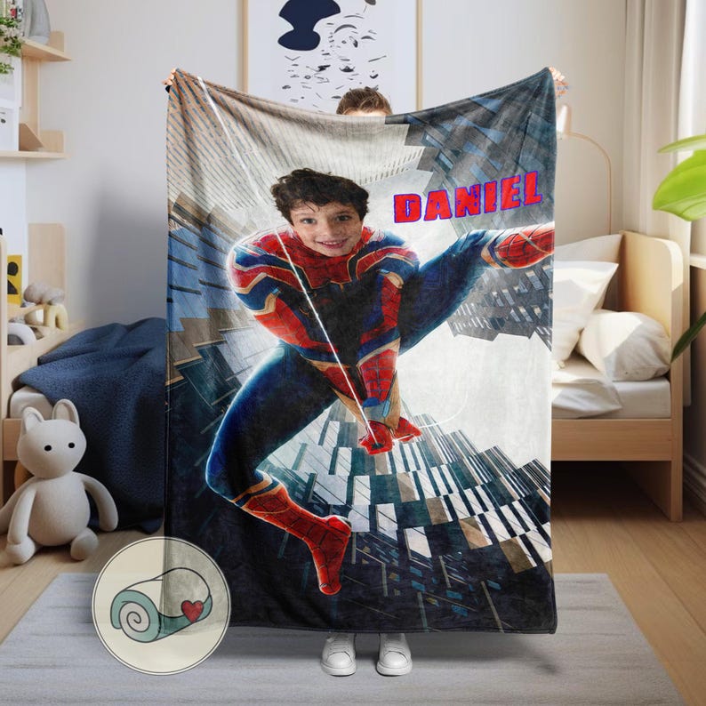 Op de afbeelding: Een gepersonaliseerde deken met een jongeman verkleed als Spider-Man, met de naam "DANIEL" in rode letters. Het ontwerp bevat een stadsgezicht achtergrond en het personage in een dynamische pose, met rode, blauwe en gouden kleuren.