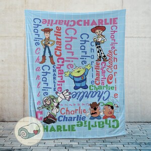 May include: A light blue blanket featuring cartoon characters and the name "Charlie" in various colors and fonts. The blanket showcases characters like Woody, Jessie, Buzz Lightyear, and others from a popular animated movie.