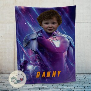 Op de afbeelding: Een gepersonaliseerde pluche deken met het gezicht van een kind bovenop een Iron Man-pakontwerp. De deken is overwegend paars met oranje tekst die "DANNY" luidt. Het ontwerp bevat een superheldenthema.
