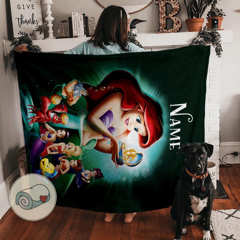 May include: A dark green blanket featuring a vibrant illustration of Ariel from The Little Mermaid, with her red hair and a blue and gold object. The blanket also includes the word "NAME" in white, and a rolled-up blanket graphic.