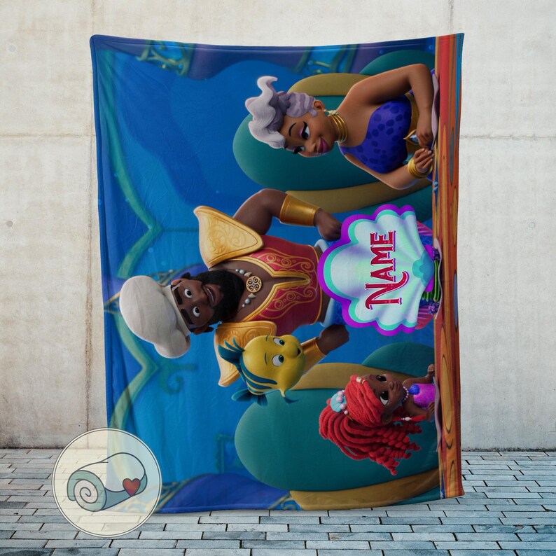 May include: A blue fleece blanket with cartoon characters from a popular animated movie. The blanket features a mermaid, a sea creature, and a man in a red and gold outfit. The word "NAME" is visible on the blanket.