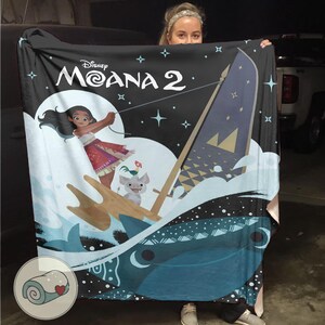 May include: A black blanket featuring the Disney movie "Moana 2" with a graphic of the main character, a boat, and ocean waves. The blanket has a rolled-up image with a heart.