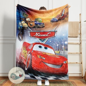 May include: A red Cars-themed blanket featuring Lightning McQueen racing on a track. The blanket showcases other characters and the text "Name" on a shield. The design includes a checkered flag pattern at the bottom.