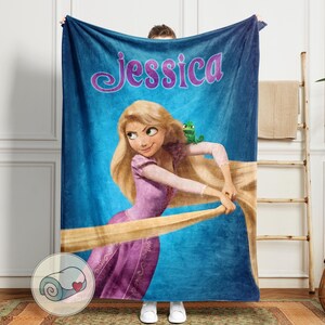May include: A blue fleece blanket featuring a cartoon illustration of a princess with long blonde hair and a green frog. The name "Jessica" is written in purple script across the top. The blanket is held up by a person.
