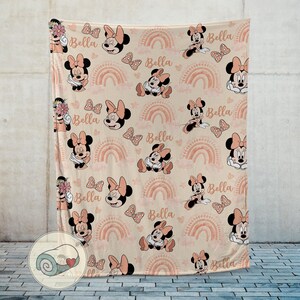 May include: A beige blanket featuring a repeating pattern of Minnie Mouse, bows, rainbows, and the name "Bella" in a soft pink color. The blanket has a rolled-up image in the corner.