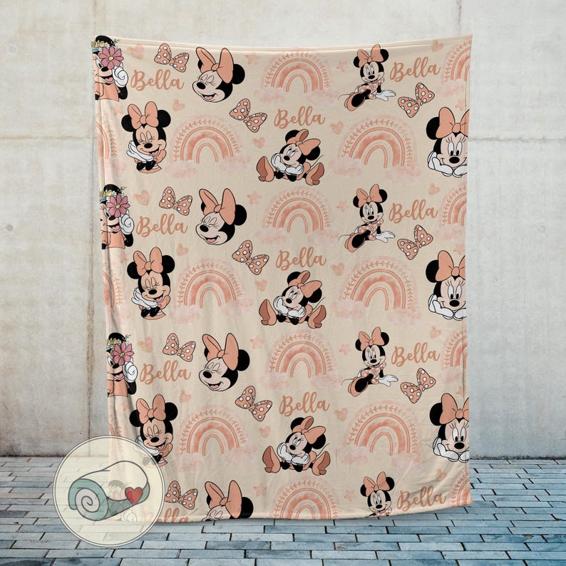 May include: A beige blanket featuring a repeating pattern of Minnie Mouse, bows, rainbows, and the name "Bella" in a soft pink color. The blanket has a rolled-up image in the corner.