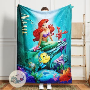 Puede incluir: Una manta turquesa con una colorida ilustraci&oacute;n de Ariel la sirenita, con su pelo rojo y su top de conchas moradas, y otros personajes de la pel&iacute;cula. La palabra "NAME" est&aacute; impresa verticalmente en el lado izquierdo.