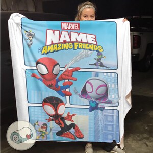 May include: A Marvel "Amazing Friends" blanket featuring Spider-Man, Spider-Gwen, and Miles Morales. The blanket has a light blue background with comic book style panels and character illustrations. The Marvel logo and "Name" text are displayed at the top.