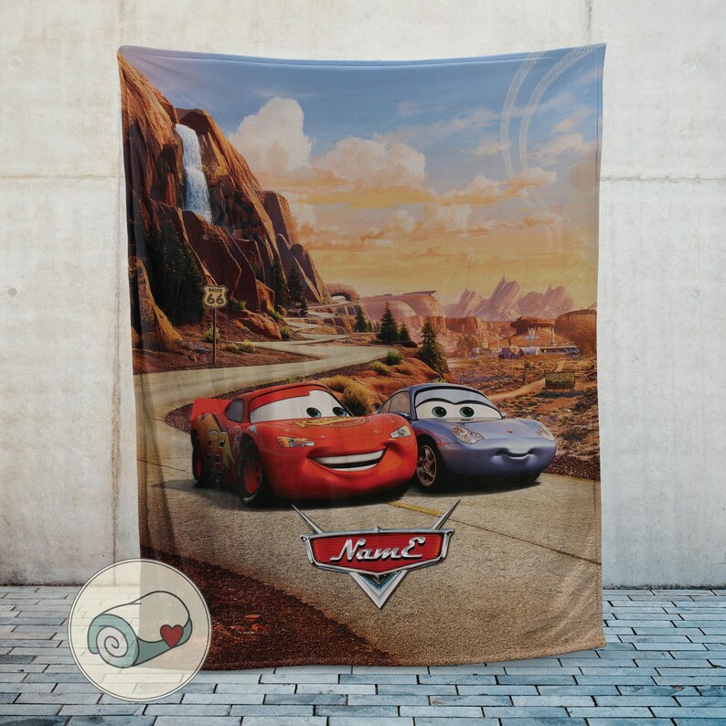 May include: A plush blanket featuring a scenic desert landscape with two animated cars, one red and one blue, on a road. The blanket has a Route 66 sign and the word "Name" in a stylized logo.