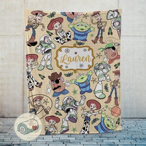 May include: A beige blanket featuring cartoon characters from a popular animated movie. The blanket has a repeating pattern of characters like Woody, Buzz Lightyear, and others. The name "Lauren" is displayed in a decorative frame.