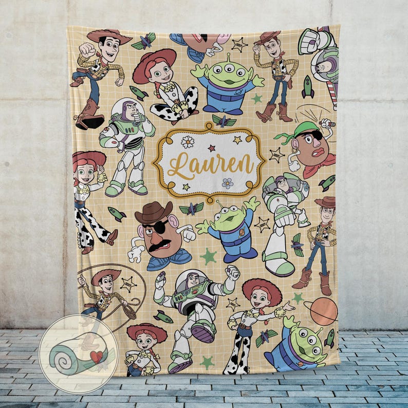 May include: A beige blanket featuring cartoon characters from a popular animated movie. The blanket has a repeating pattern of characters like Woody, Buzz Lightyear, and others. The name "Lauren" is displayed in a decorative frame.