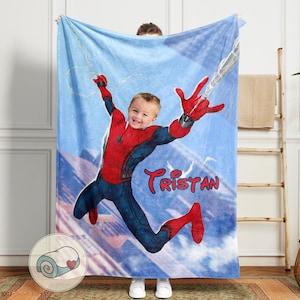 Op de afbeelding: Een blauwe deken met een gepersonaliseerde afbeelding van een kind verkleed als Spider-Man, met de naam "Tristan" in rood schrift. De deken heeft een lichtblauwe hemelachtergrond met een webontwerp.