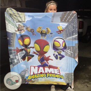 May include: A colorful blanket featuring cartoon characters, including Spider-Man and other heroes, against a cityscape backdrop. The blanket has the text "NAME" and "THE AMAZING FRIENDS" in a bold font. The blanket is a vibrant mix of blues, reds, and yellows.