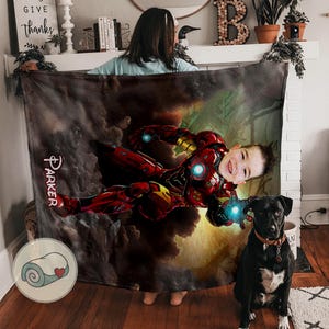 May include: A personalized blanket featuring a child in an Iron Man suit, set against a cloudy background. The name "Parker" is written on the blanket. A black dog with a brown collar sits in front of the blanket.