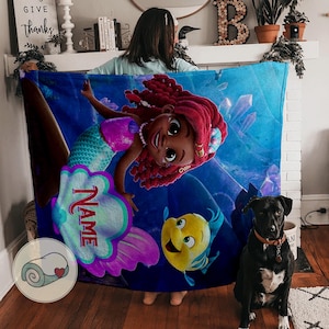 Peut inclure: Une couverture bleue avec une sir&egrave;ne de dessin anim&eacute; aux cheveux roux, un haut rose et une queue turquoise. La couverture a une zone de nom personnalis&eacute;e et un poisson jaune. Un chien noir est devant.