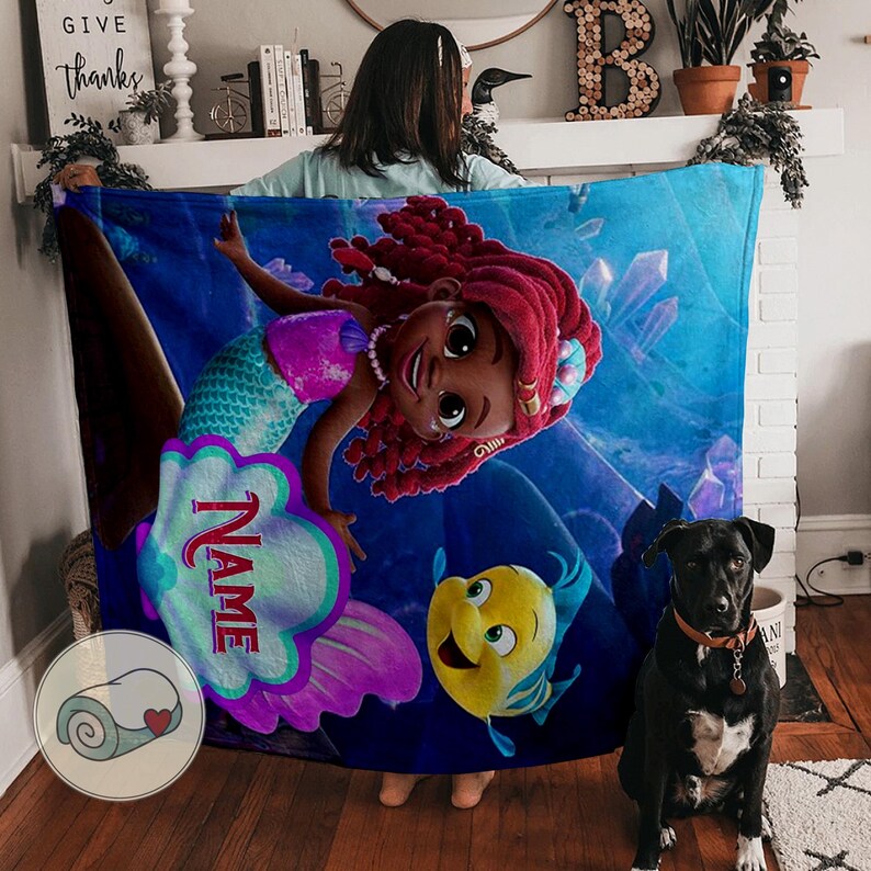 Peut inclure: Une couverture bleue avec une sir&egrave;ne de dessin anim&eacute; aux cheveux roux, un haut rose et une queue turquoise. La couverture a une zone de nom personnalis&eacute;e et un poisson jaune. Un chien noir est devant.
