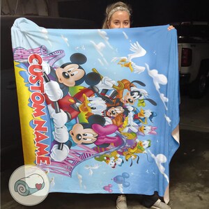 May include: A blue blanket featuring Mickey Mouse and friends, with a custom name in red and white. The blanket has a cartoon design with clouds and birds, and a small image of a rolled-up blanket with a heart.