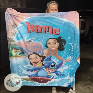 May include: A blue and white blanket featuring a cartoon scene with the characters Lilo, Nani, and Stitch surfing a wave. The word "Name" is printed in red across the top. The blanket has a beach scene background.