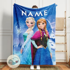 May include: A blue fleece blanket featuring Elsa and Anna from Frozen, with the word "NAME" in white at the top. The blanket has a wintery background with snowflakes and a mountain. The blanket is held up by a person.