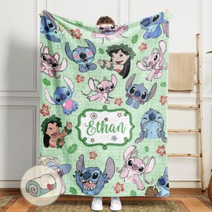 Puede incluir: Una manta verde claro con varios personajes de dibujos animados en azul y rosa, junto con detalles florales rojos y rosas. La manta tiene un patr&oacute;n de cuadr&iacute;cula y una etiqueta central con el nombre "Ethan" en verde.