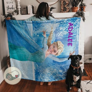 May include: A blue and white blanket featuring a graphic of a smiling blonde girl in a blue dress. The name "ADALEE" is printed vertically in pink. A black dog with a brown collar sits in front of the blanket.