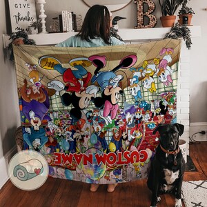 Pu&ograve; includere: Una coperta beige con un colorato design a cartoni animati con Topolino, Minnie e altri personaggi Disney. La coperta ha la scritta "CUSTOM NAME" in basso. Un cane nero con un collare marrone &egrave; seduto davanti alla coperta.
