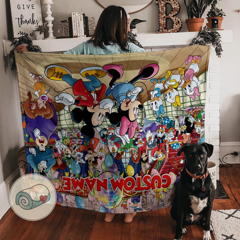 Pu&ograve; includere: Una coperta beige con un colorato design a cartoni animati con Topolino, Minnie e altri personaggi Disney. La coperta ha la scritta "CUSTOM NAME" in basso. Un cane nero con un collare marrone &egrave; seduto davanti alla coperta.