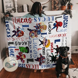 Puede incluir: Manta blanca con ilustraciones de superh&eacute;roes y el nombre "Lucas" en rojo y azul. La manta tiene un patr&oacute;n repetido del nombre y personajes de superh&eacute;roes. Un perro negro est&aacute; sentado frente a la manta.