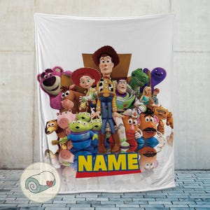 May include: White blanket featuring a colourful group of Toy Story characters, including Woody, Buzz Lightyear, and Mr. Potato Head. The word "NAME" is printed in yellow with a red border at the bottom. The blanket is displayed against a light grey wall.